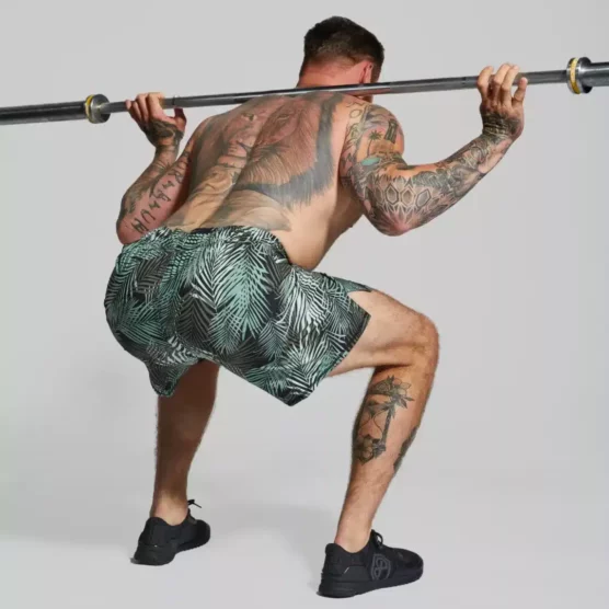 Mann i grønn og sort herreshorts i teknisk stoff som står i en squat med ryggen mot kameraet med en barebell på nakken. Shortsen har palmebladmotiv i grønn farge.