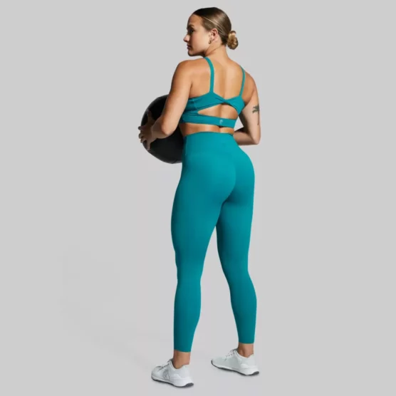 Dame i grønn tights og sports-bh i teknisk stoff som står med ryggen mot kameraet og holder en medisinball foran seg.. Tightsen har høyt liv.