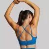 Dame i blå sports-bh i teknisk stoff med ryggen mot kameraet og strekker armene over hodet. Sports-bhen har en stropp over hver skulder, samt to stropper som krysser hverandre bak.