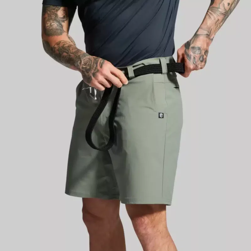 Beina til en mann i grønn shorts i teknisk stoff vendt skrått med fronten mot kameraet. Mannen tar belte på shortsen.