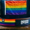 Løftebelte fra merket 2Pood Performance som ligger på et bord. Beltet har striper i Pride-fargene. Borrelåsen er sort og det står 2POOD med hvit skrift.