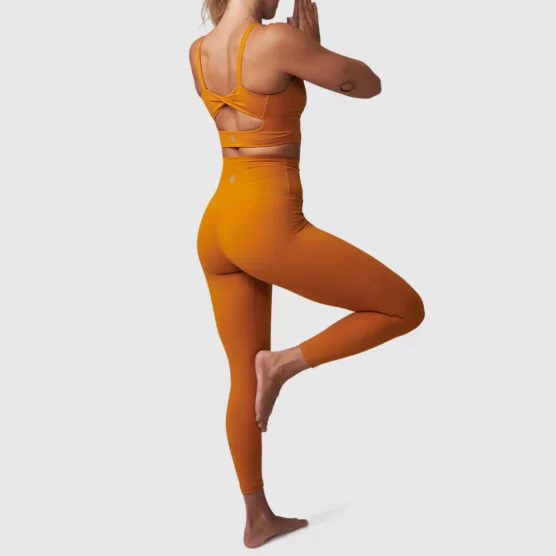 Dame i oransje tights i teknisk stoff som står med ryggen skrått mot kameraet i en yogapositur. Tightsen er høy i livet.
