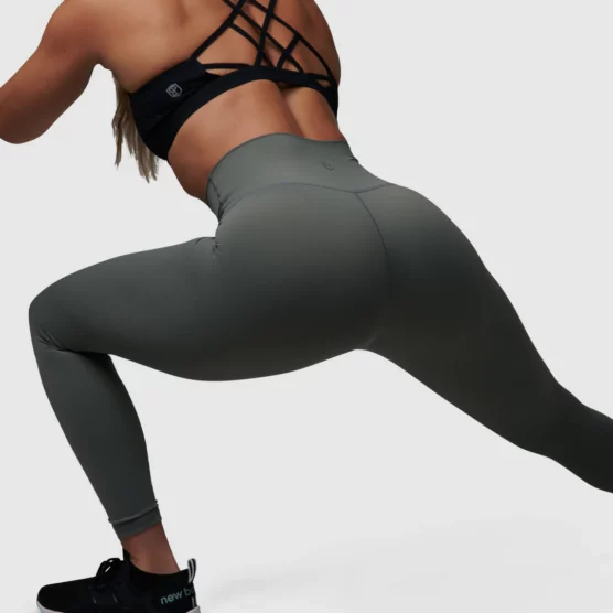 Dame i grå tights i teknisk stoff som står i en stretchingposisjon med ryggen skrått mot kameraet. Tightsen har høyt liv.