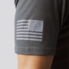 Armen til en mann i grå t-skjorte som står med siden mot kameraet. På ermet er det bilde av det amerikanske flagget i hvitt.