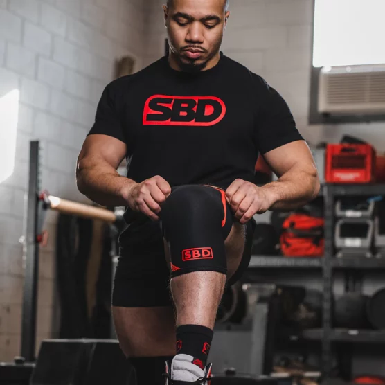 Mann som står vendt mot kameraet og tar på knee sleeves fra SBD. Knee sleevsene er sorte og det står SBD i en rød firkant i rødt nederst foran.