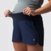 Beina til en gravid dame i blå shorts i teknisk stoff som står skrått vendt mot kameraet. Shortsen har et elastisk og bredt magebelte.
