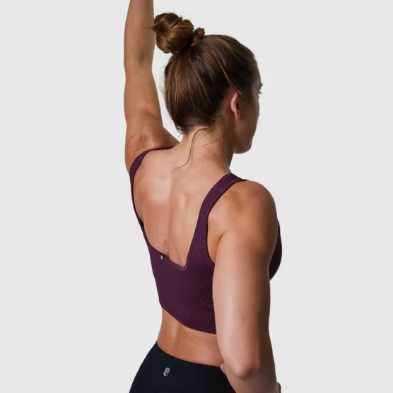 Dame i kirsebærfarget sports-bh med ryggen skrått mot kameraet. Den har brede stropper over skuldrene og et bredt bånd nederst. Sports-bhen er ribbestrikket.