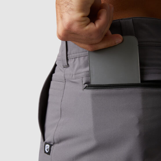 Beina til en mann i mørkegrå shorts i polyester. Bildet viser utsnitt av lommen bak på den venstre siden og mannen putter en telefon i lommen. Lommen har en sort glidelås.