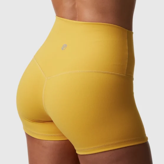 Beina til en dame i sennepsgul shorts i teknisk stoff som står med ryggen mot kameraet. Shortsen er høy i livet.