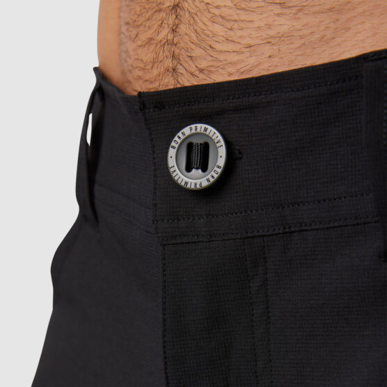 Beina til en mann i sort bukse i teknisk stoff vendt med fronten mot kameraet. Den har glidelås og knapp, samt hemper til belte.