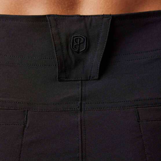 Korsryggen til en mann i sort bukse i teknisk stoff med ryggen mot kameraet. Bildet viser et utsnitt av en hempe til belte midt på bak. Det er en brodert Born Primitive-logo på hempen.