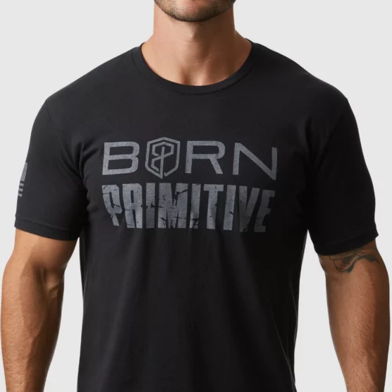 Mann i sort t-skjorte i teknisk stoff som står vendt mot kameraet. Det står "Born Primitive" med grå bokstaver på brystet. t-skjorte
