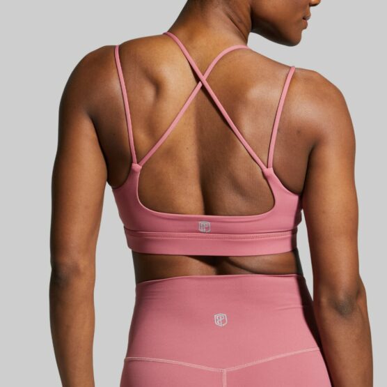 Dame i rosa sports-bh og shorts i teknisk stoff med ryggen mot kameraet. Sports-bhen har en stropp over hver skulder, samt to stropper som krysser hverandre bak.