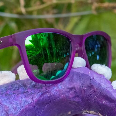 Lilla solbriller med reflekterende glass fra Goodr som ligger oppå en lilla hale med hvite bomullsdotter.. Speilbildet i glassene viser grønn vegetasjon. På siden av innfatningen står det goodr.
