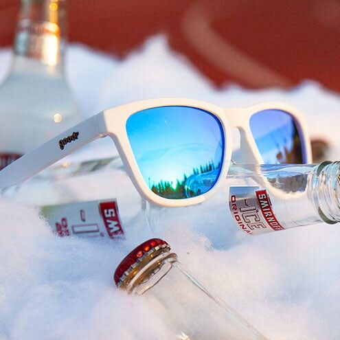Hvite solbriller med reflekterende glass fra Goodr. Speilbildet i glassene viser blå himmel og trær. Brillene ligger oppå en Smirnoff Ice-flaske som ligger på is. På siden av innfatningen står det goodr.