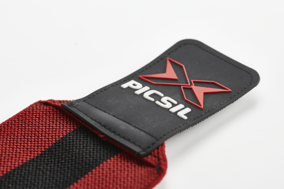 Oversiden av borrelåsen på en sort og rød wrist wrap fra Picsil. Det er en rød X og det står Picsil med hvit skrift.