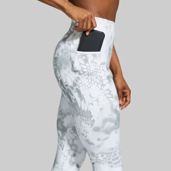 Beina til en dame som står med siden vendt mot kameraet og putter telefonen i en sidelomme på tightsen. Hun har på seg en Eccentric legging som er lys grå og hvitmønstret.