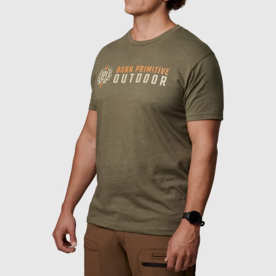 Mann i ført militærgrønn T-skjorte avbildet skrått forfra, med skriften Born Primitive i orange, og Outdoor i hvitt. På brystet. Det også en loge ved siden av.