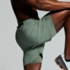 Mann avbildet fra livet og ned iført en grønn shorts, der han står på et ben, mens han løfter og holder hendene på det andre kneet.