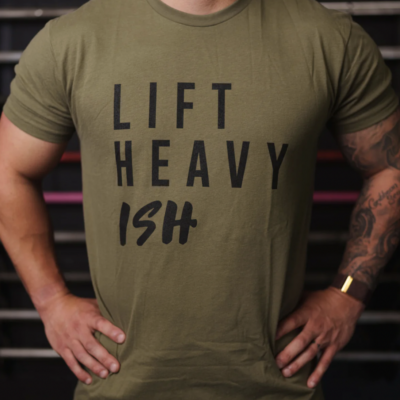 Lift Heavy Ish T-shirt, Overkroppen til en mann som står med hendene i siden, iført en grønn t-skjorte med følgende teks på brystet; Lift heavy Ish