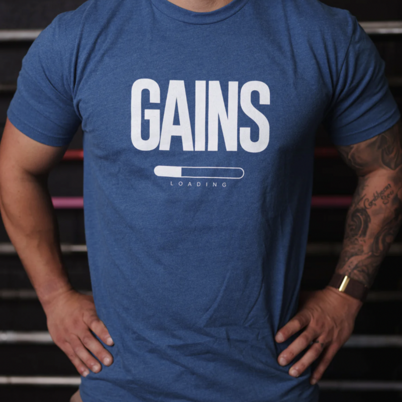 gains Loading T-shirt Overkroppen til en mann som står med hendene i siden, iført en kongeblå t-skjorte med følgende tekst på brystet; Gains