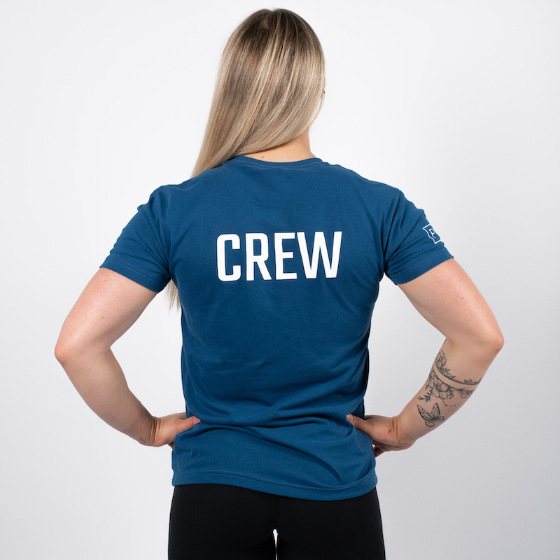 Kvinne avbildet bakfra med armene i siden, iført en blå t-skjorte med skriften CREW skrevet i hvit midt bak på ryggen.