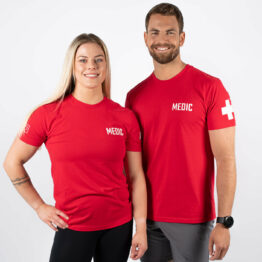 unisex tskjorte, medic tskjorte. Mann og kvinne avbildet forfra iført rød tskjorte med skriften MEDIC skrevet i hvit på venstre bryst, og med hvit "førstehjelps-merke" på venstre overarm.
