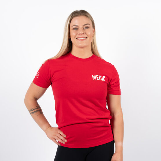 Kvinne avbildet forfra med en arm i siden. iført en rød t-skjorte med skriften MEDIC skrevet i hvit på venstre bryst.