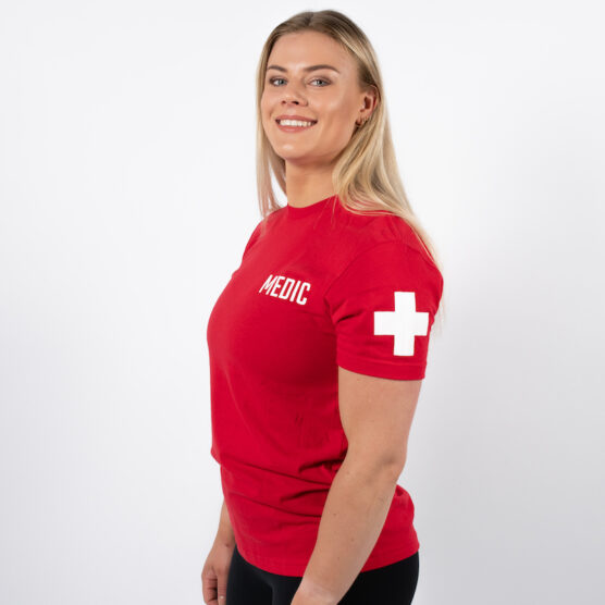 Kvinne avbildet skrått forfra iført en rød t-skjorte med skriften MEDIC skrevet i hvit på venstre bryst og et hvit "førstehjelps-kryss" på venstre overarm.