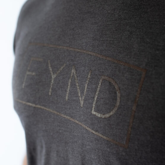 Detaljbilde av kaffebrun t-skjorte med teksten FYND skrevet i et rektangel med brun skrift.