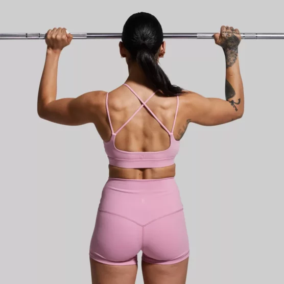 En kvinne som står med ryggen mot kameraet. Hun har på seg Exhale rosa sports-bh og rosa New Heights shorts. Sports-bh'en har to stropper på hver side. To av stroppene krysser hverandre bak på ryggen. Hun trener med en vektstang.