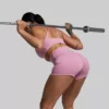 En kvinne som står med ryggen skrått vendt mot kameraet og har på seg en rosa New Heights shorts og Exhale rosa sports-bh. Hun trener med en vektstang.