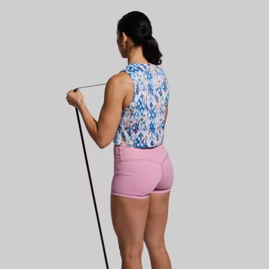 Kvinne som trener med strikk og står med ryggen mot kameraet. Hun har på seg en Subtle flex tank topp og en rosa New Heights shorts. Toppen har et mønster i blå, hvit, rosa, sort, brun og gul.