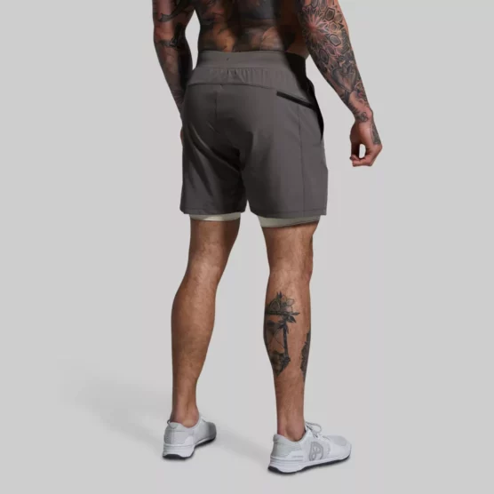 Beina til en mann som står med ryggen mot kameraet. Han har på seg en grå Versatile shorts. Shortsen har lommer foran og en lomme med glidelås bak på den høyre siden. Den har en innebygd kremhvit kompresjonsshorts.