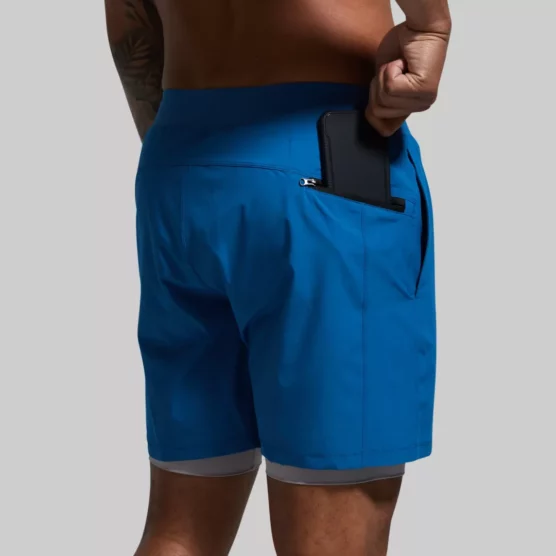 Beina til en mann som står med ryggen mot kameraet. Han har på seg en blå Versatile shorts. Shortsen har lommer foran og en lomme med glidelås bak på den høyre siden. Den har en innebygd grå kompresjonsshorts.