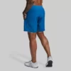 Beina til en mann som står med ryggen mot kameraet. Han har på seg en blå Versatile shorts. Shortsen har lommer og den har en innebygd grå kompresjonsshorts.