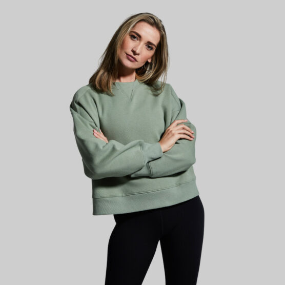 Pump Sweatshirt (Aspen) Kvinne står vendt mot kamera med armene i kryss. Hun har på seg en dus grønn Pump Sweatshirt og svarte tights.